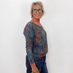 A woman with short blonde hair and glasses, wearing a long-sleeve knit top with a blue, teal, and rust-orange abstract pattern, paired with dark blue jeans and gold bracelets, standing against a plain white wall.