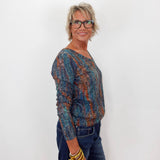A woman with short blonde hair and glasses, wearing a long-sleeve knit top with a blue, teal, and rust-orange abstract pattern, paired with dark blue jeans and gold bracelets, standing against a plain white wall.