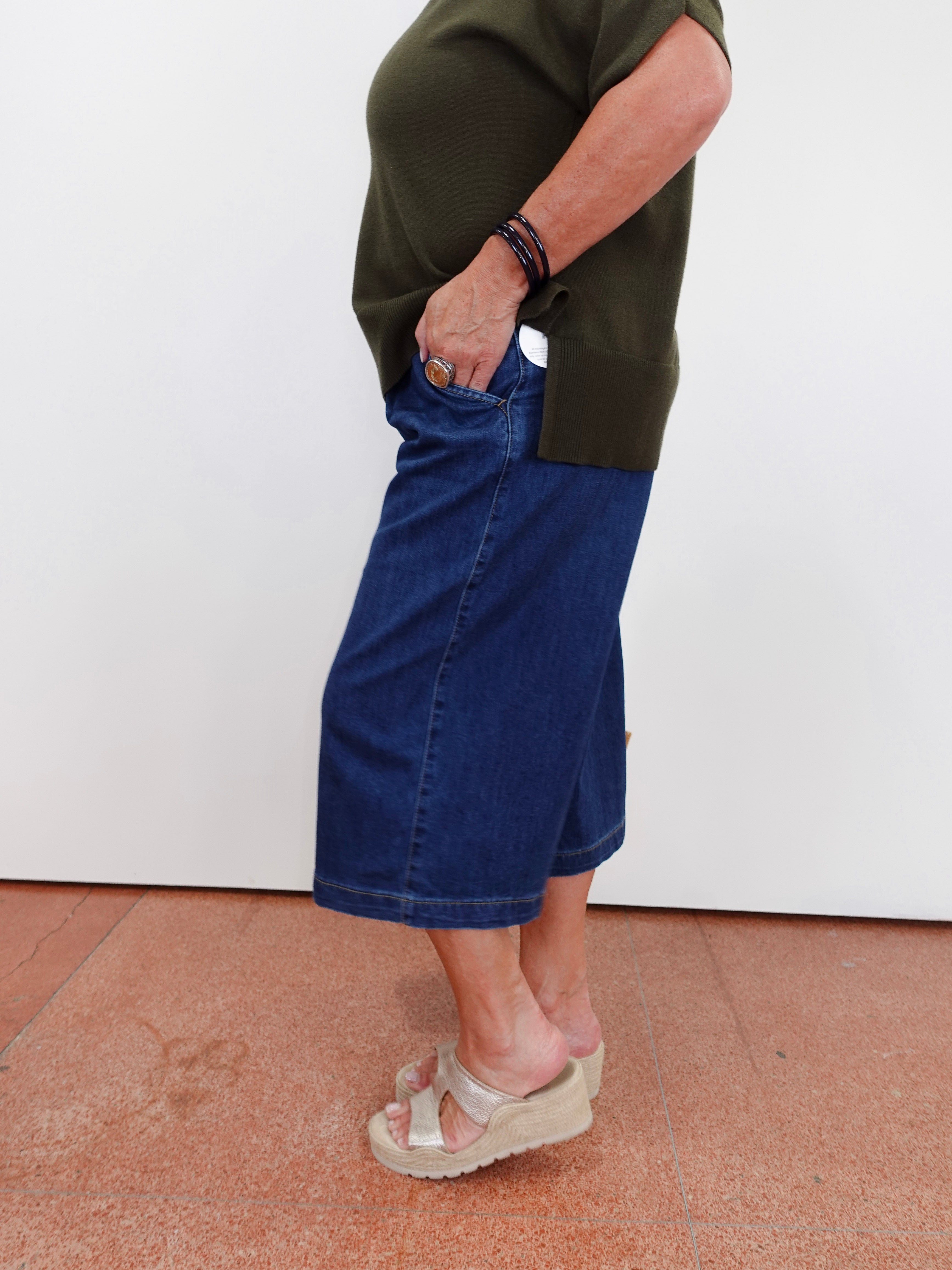 A person wearing a dark blue denim midi skirt, an olive green top, and beige platform sandals, standing against a white wall.