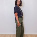 Woman wearing a dark blue top and green pants against a white wall.