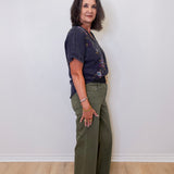 Woman wearing a dark blue top and green pants against a white wall.