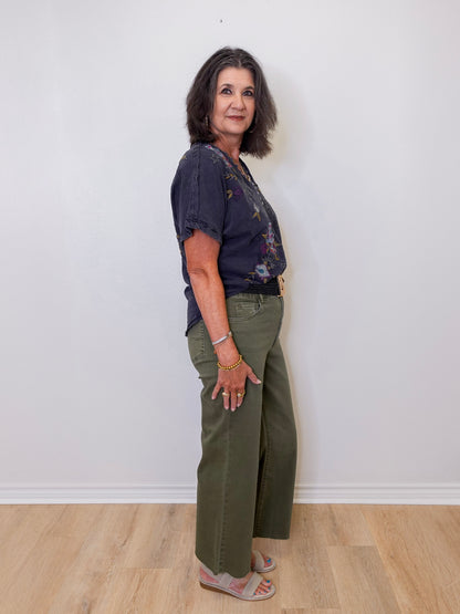 Woman wearing a dark blue top and green pants against a white wall.