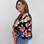 A woman wearing a black floral blouse with orange, pink, blue, and yellow flowers, paired with light blue jeans, standing against a plain light gray background.