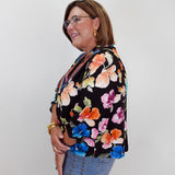 A woman wearing a black floral blouse with orange, pink, blue, and yellow flowers, paired with light blue jeans, standing against a plain light gray background.