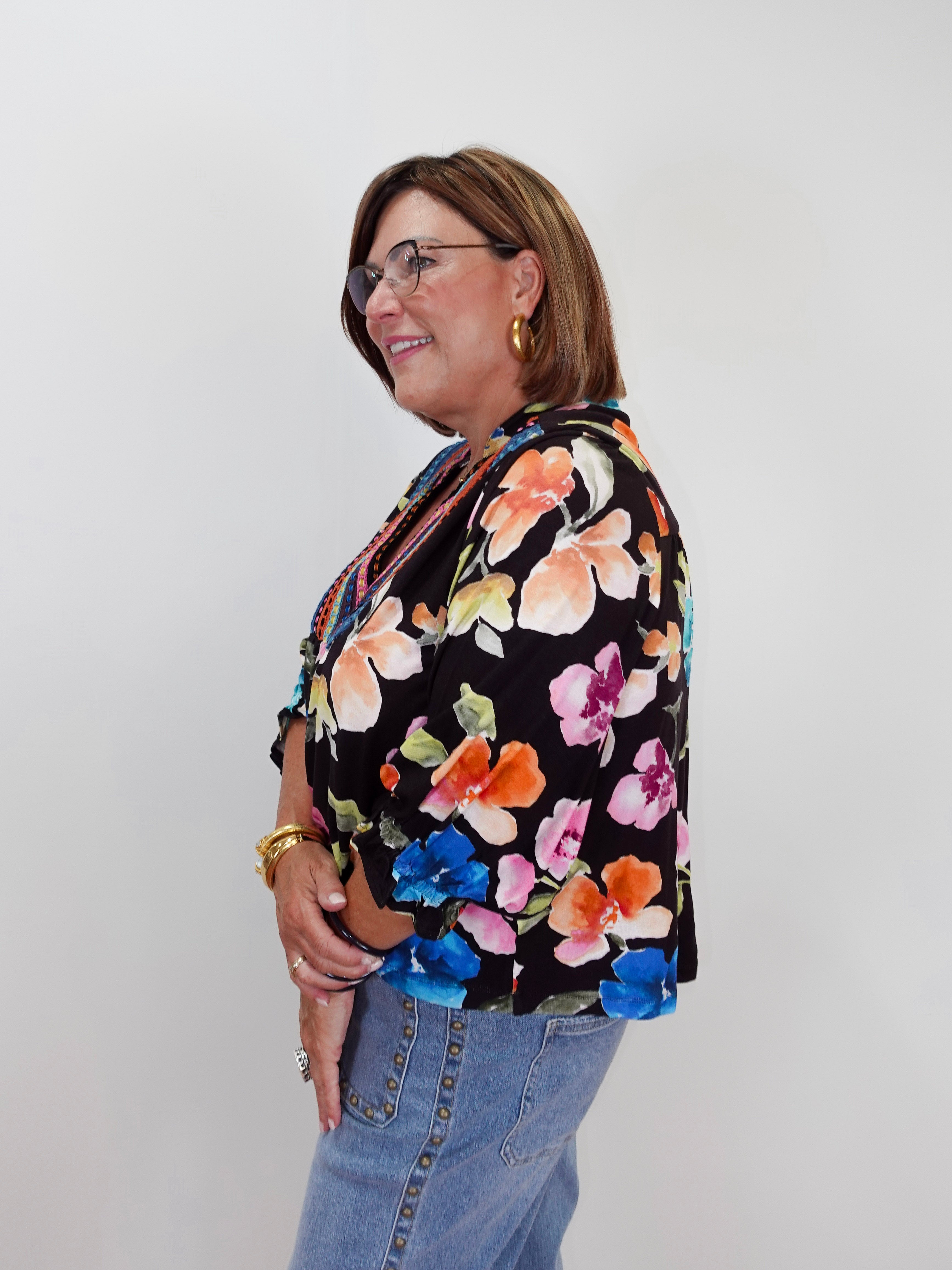 A woman wearing a black floral blouse with orange, pink, blue, and yellow flowers, paired with light blue jeans, standing against a plain light gray background.