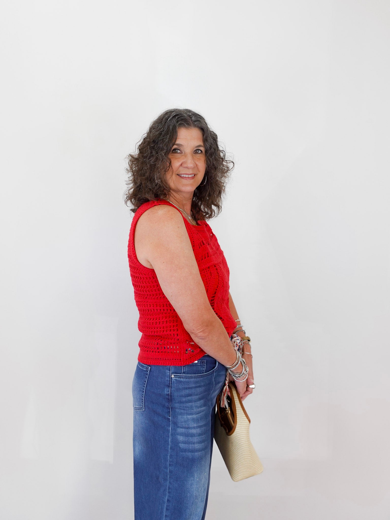 Woman wearing a red sleeveless top and blue jeans, holding a beige handbag against a white background