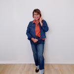 Person wearing a denim jacket and jeans standing against a white wall.