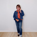 Person wearing a denim jacket and jeans standing against a white wall.