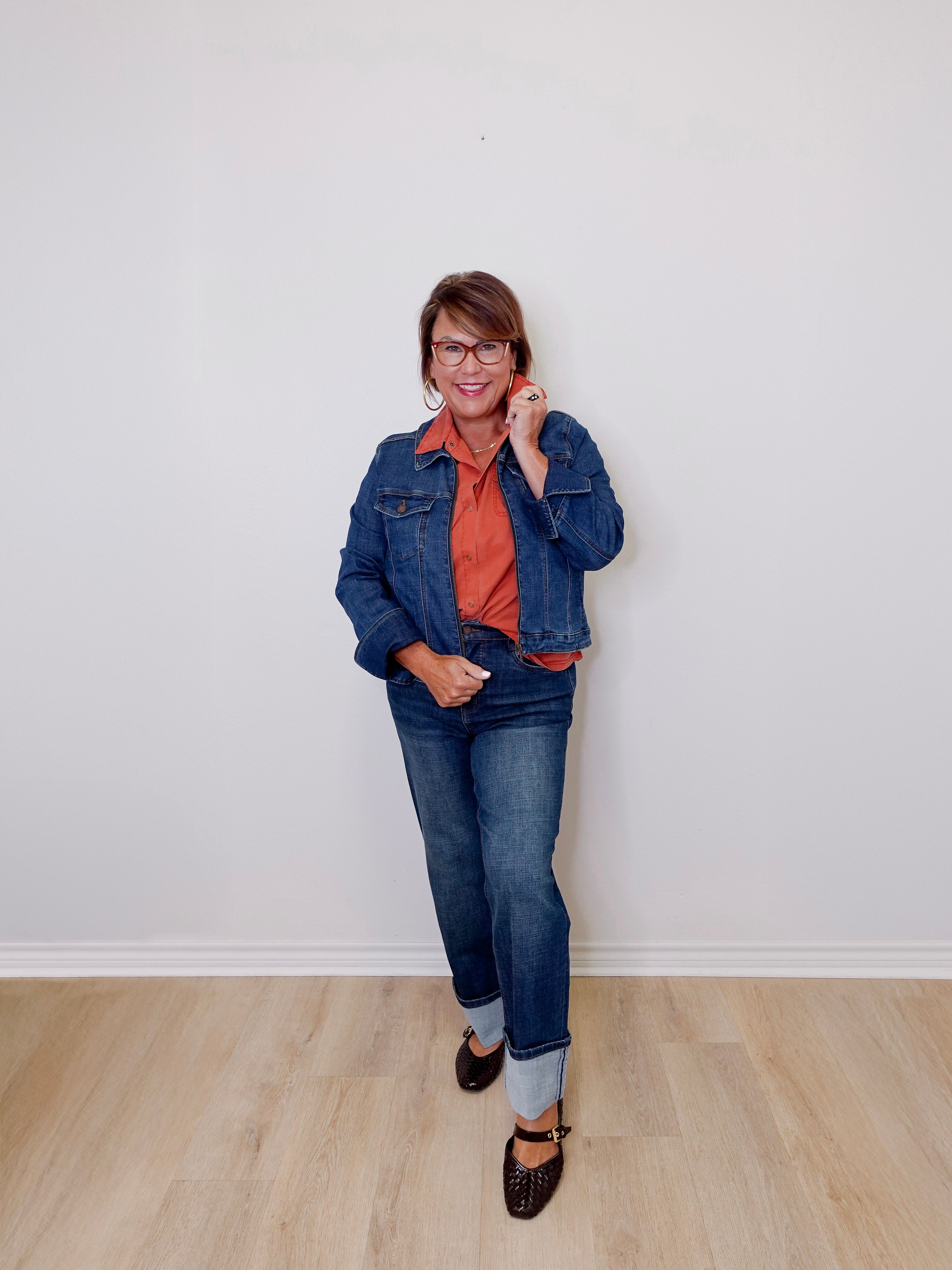 Person wearing a denim jacket and jeans standing against a white wall.