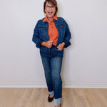 Person wearing a blue denim jacket, orange shirt, and jeans standing against a white wall.