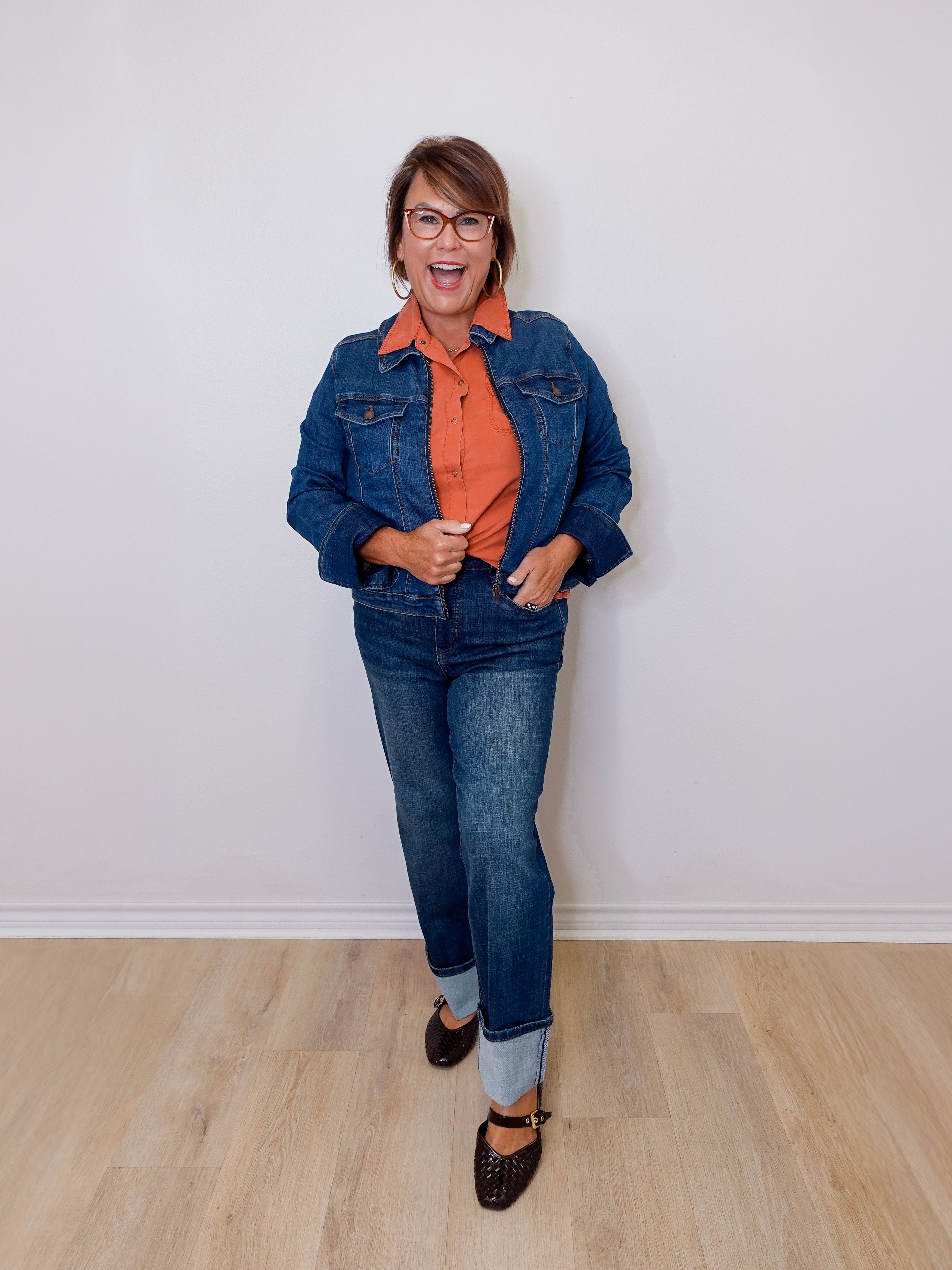 Person wearing a blue denim jacket, orange shirt, and jeans standing against a white wall.