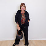 Woman in a black outfit with a patterned top, holding a handbag against a white wall.