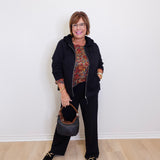 Woman in a black outfit with a patterned top, holding a handbag against a white wall.