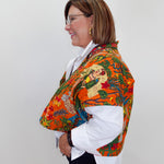 A woman shown in profile wearing a vibrant orange shawl with a detailed Mexican folk art print over a white collared shirt and dark pants, set against a plain light gray background.
