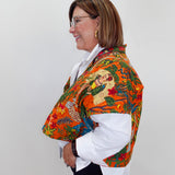 A woman shown in profile wearing a vibrant orange shawl with a detailed Mexican folk art print over a white collared shirt and dark pants, set against a plain light gray background.