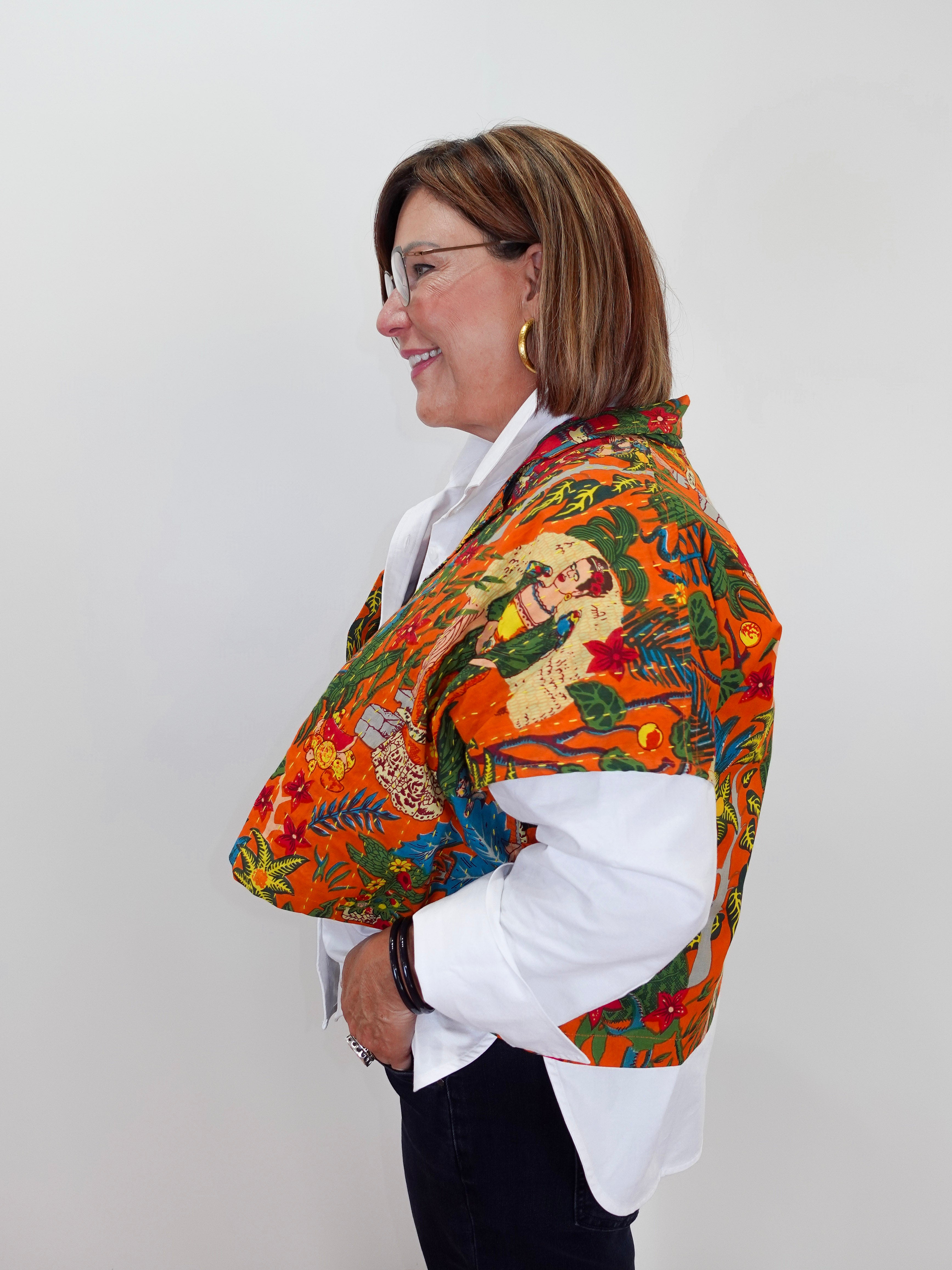 A woman shown in profile wearing a vibrant orange shawl with a detailed Mexican folk art print over a white collared shirt and dark pants, set against a plain light gray background.