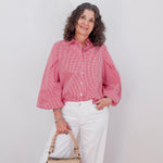 Woman wearing a pink checkered blouse and white pants, holding a beige handbag against a white background.