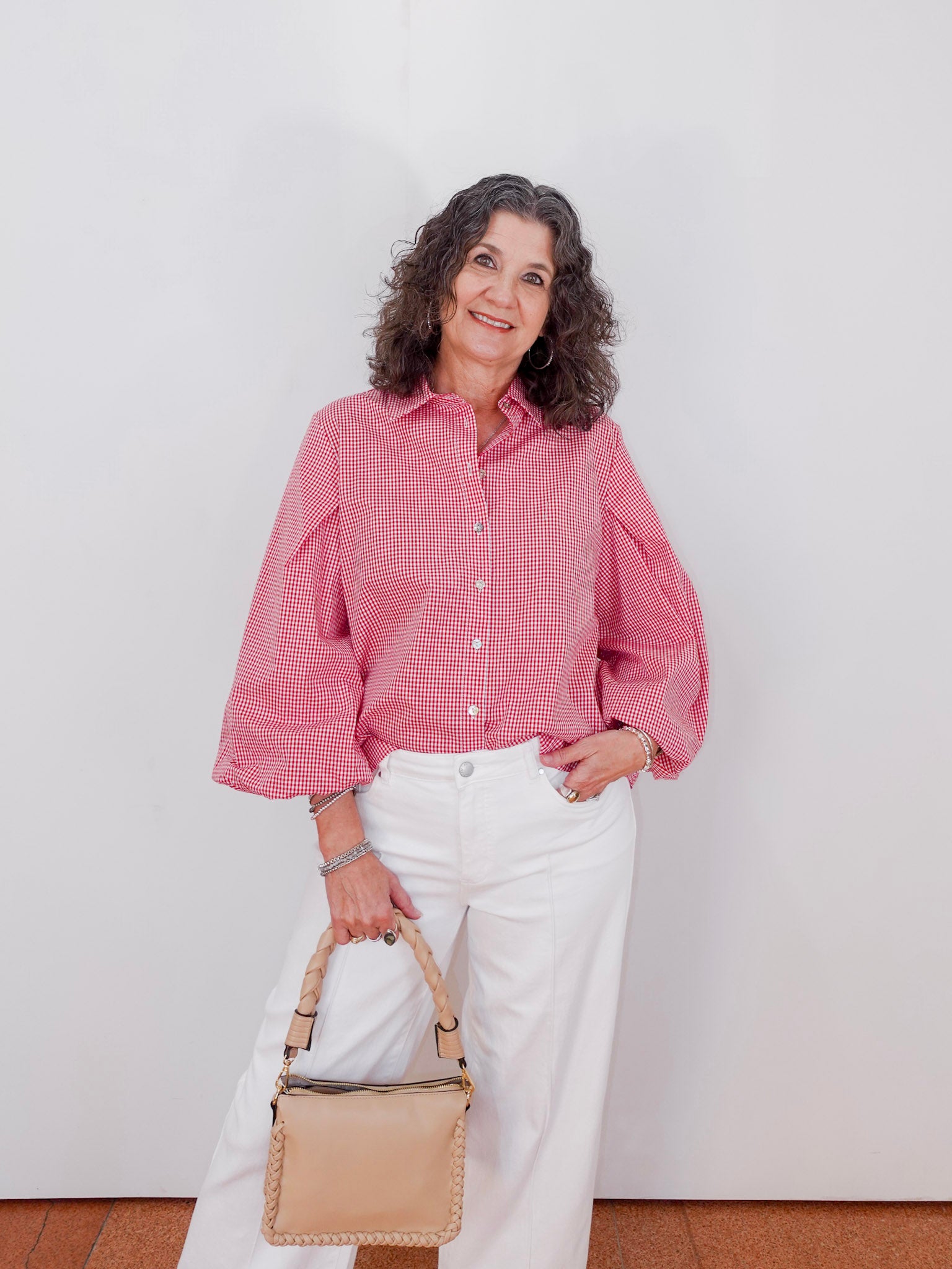 Woman wearing a pink checkered blouse and white pants, holding a beige handbag against a white background.