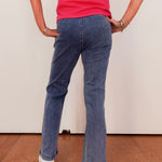 Person wearing a pink top and blue jeans standing against a white wall.