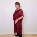 Woman wearing a burgundy dress with ruffled layers against a white wall.