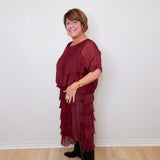 Woman wearing a burgundy dress with ruffled layers against a white wall.