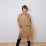 Woman wearing a beige layered dress with black boots against a white wall.