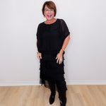 Woman wearing a black outfit with sheer layers against a white wall.