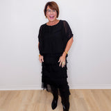 Woman wearing a black outfit with sheer layers against a white wall.