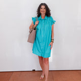 Woman wearing a turquoise dress holding a beige handbag against a white wall.