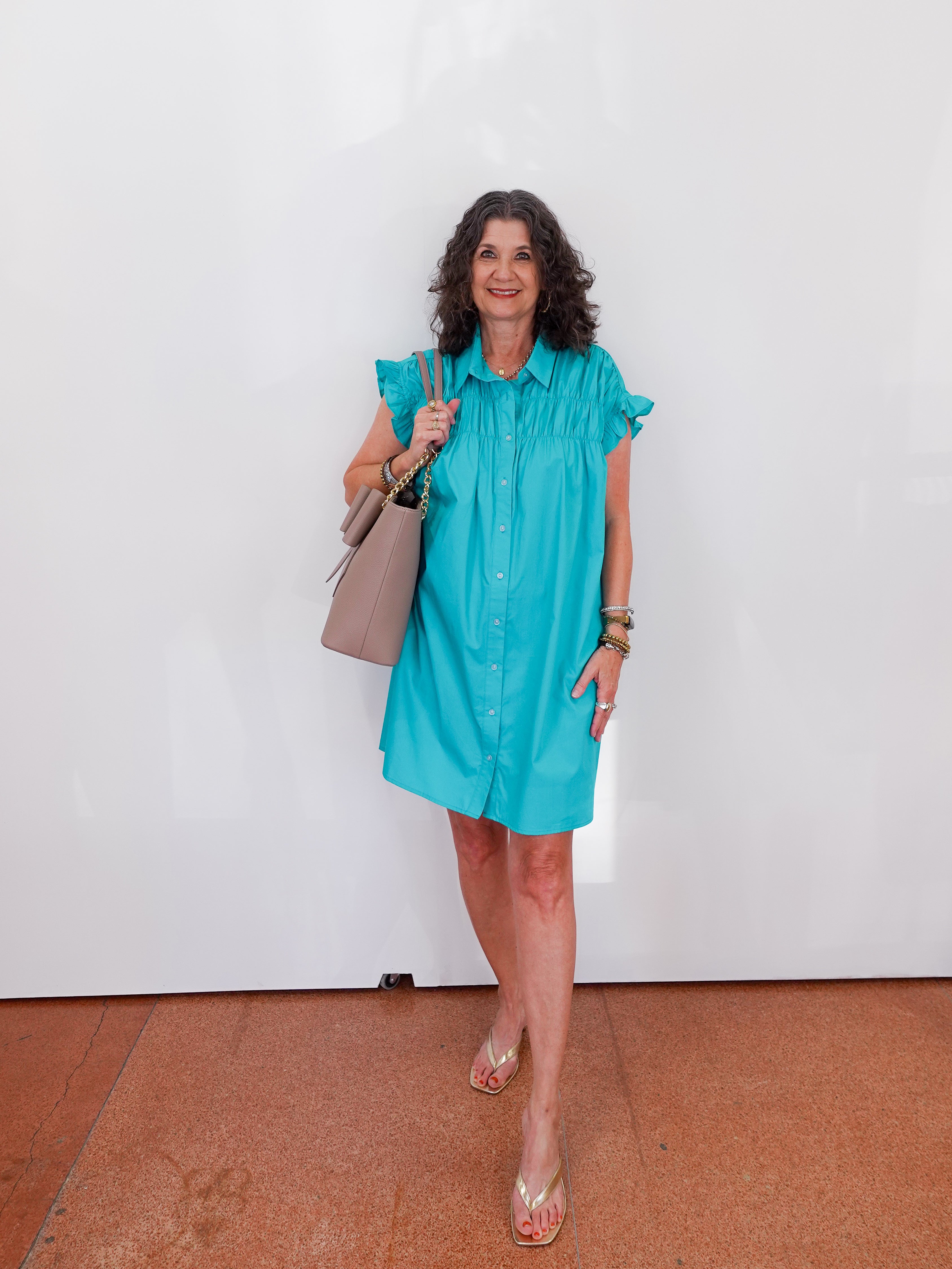 Woman wearing a turquoise dress holding a beige handbag against a white wall.