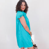 Woman wearing a turquoise dress holding a beige handbag against a white wall.