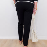 Person wearing black pants with white trim, holding a white handbag against a white wall.
