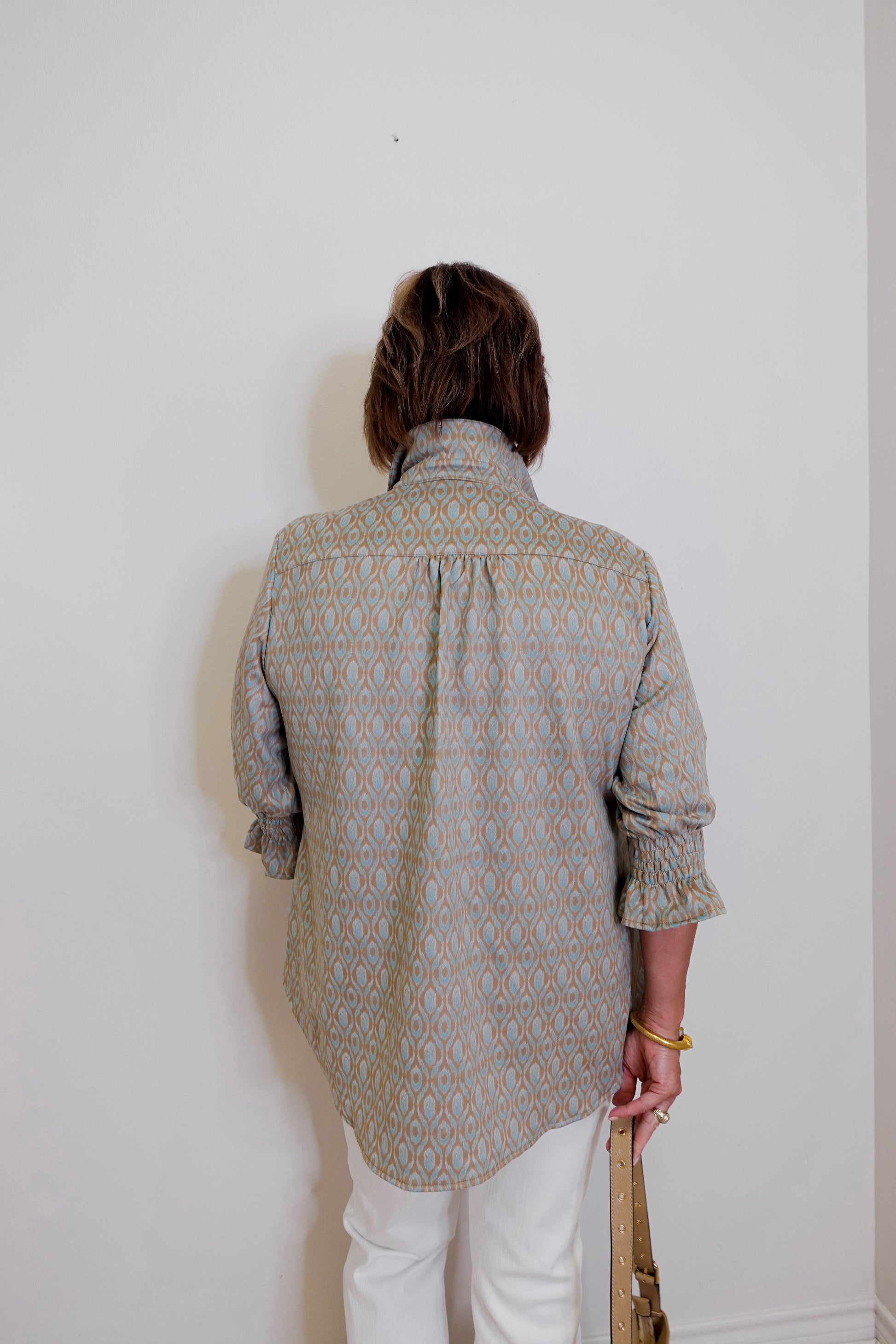 Person wearing a patterned shirt and white pants against a plain wall.