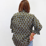 A woman viewed from behind wearing a long-sleeve button-up blouse with a green and gray geometric pattern, paired with light blue jeans, against a plain white background.