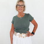 Woman wearing a green top and white pants with a gold belt against a white background