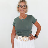 Woman wearing a green top and white pants with a gold belt against a white background