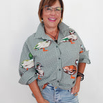 A smiling woman with glasses is wearing a green and white gingham button-up shirt with embroidered ducks on the front, paired with light blue jeans, against a plain white background.