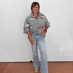 A woman wearing a gray and white gingham button-up shirt with colorful duck illustrations, light-wash blue jeans, and beige sandals, standing against a plain white wall.