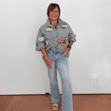 A woman wearing a gray and white gingham button-up shirt with colorful duck illustrations, light-wash blue jeans, and beige sandals, standing against a plain white wall.
