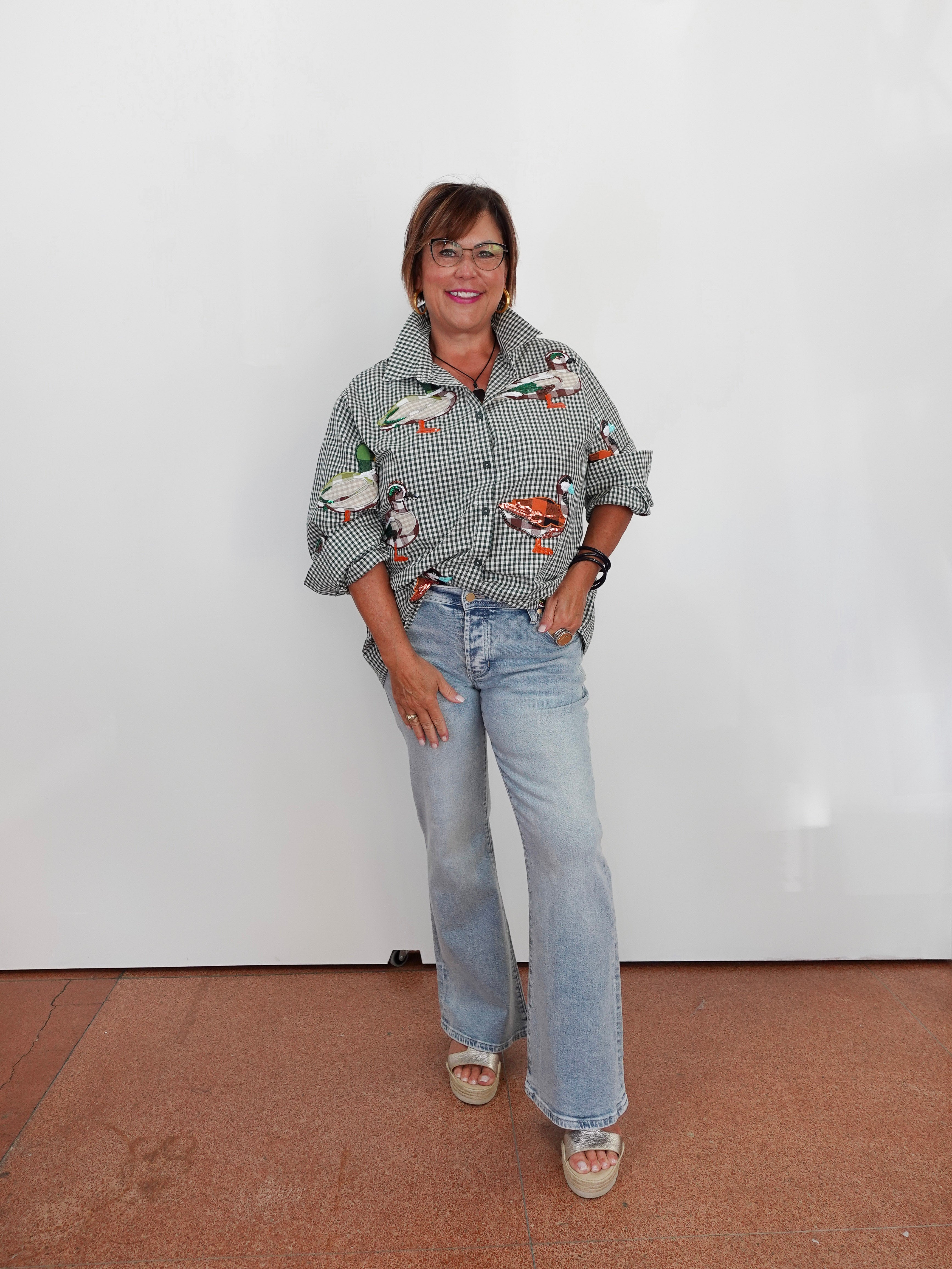 A woman wearing a gray and white gingham button-up shirt with colorful duck illustrations, light-wash blue jeans, and beige sandals, standing against a plain white wall.