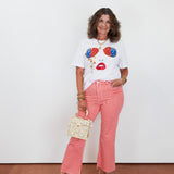 Woman wearing a white t-shirt with colorful design, red checkered pants, and holding a small handbag against a white background.