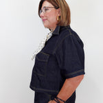 A woman with short brown hair and glasses, wearing a dark indigo denim short-sleeve shirt and matching denim pants, posing in profile against a plain white background.