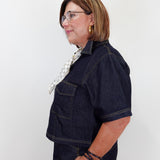 A woman with short brown hair and glasses, wearing a dark indigo denim short-sleeve shirt and matching denim pants, posing in profile against a plain white background.