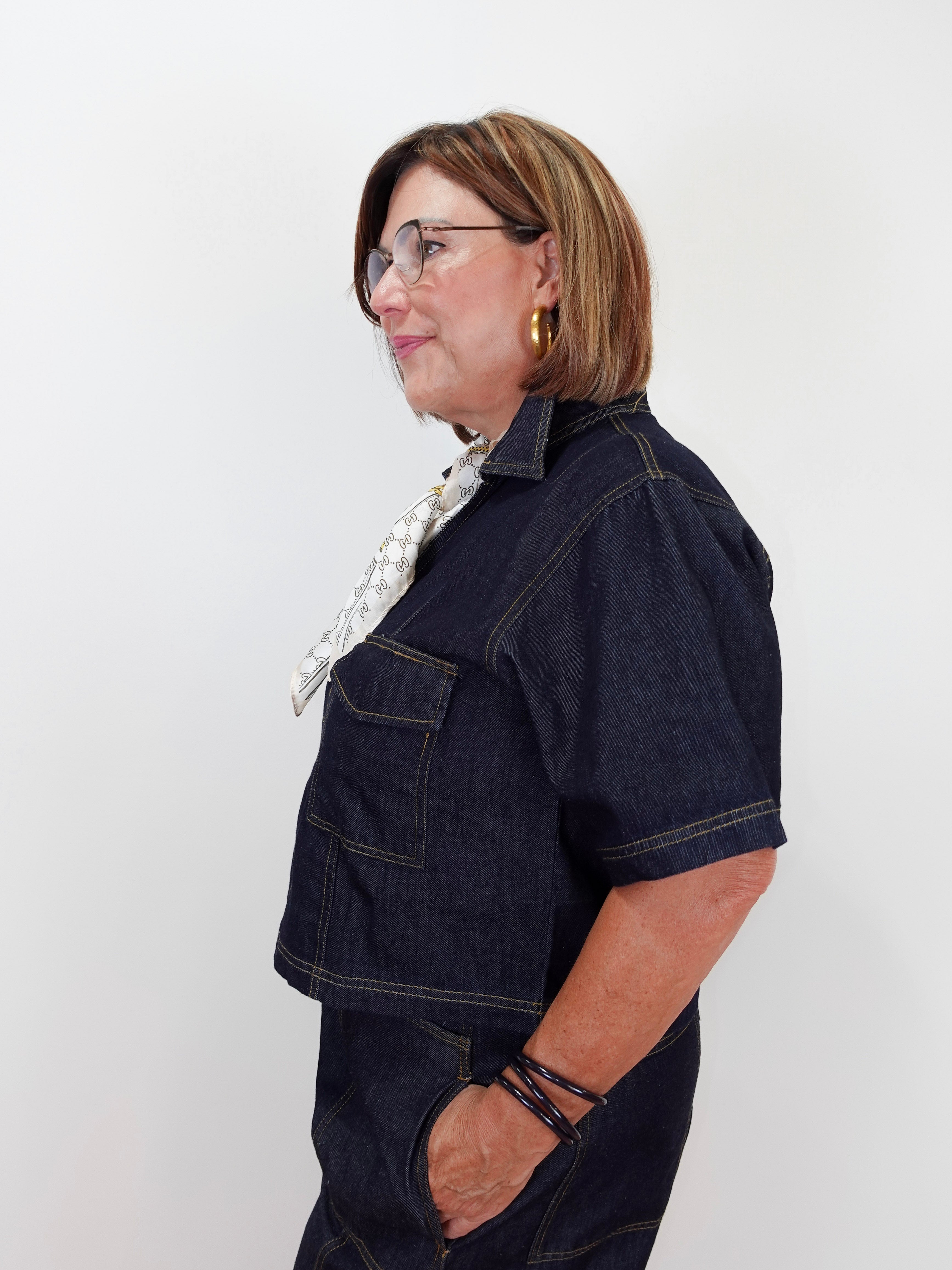 A woman with short brown hair and glasses, wearing a dark indigo denim short-sleeve shirt and matching denim pants, posing in profile against a plain white background.