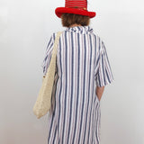 Person wearing a striped shirt and red hat with a woven bag, standing against a white wall.