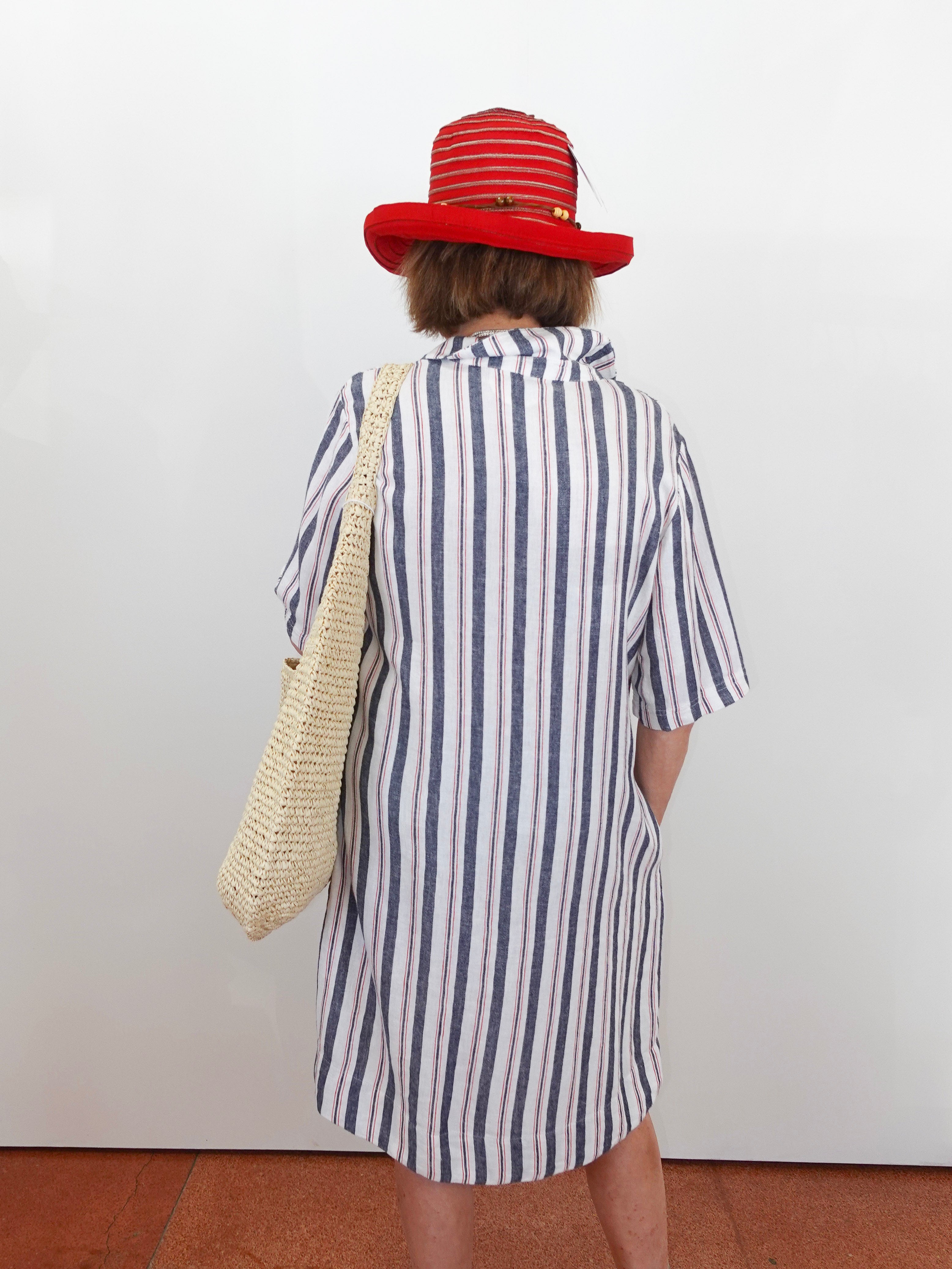 Person wearing a striped shirt and red hat with a woven bag, standing against a white wall.