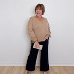 Woman wearing a beige sweater and black pants against a white wall.