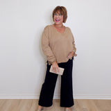 Woman wearing a beige sweater and black pants against a white wall.