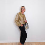 Woman wearing a gold blouse, black pants, and silver shoes against a white wall.