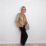 Woman wearing a gold blouse, black pants, and silver shoes against a white wall.